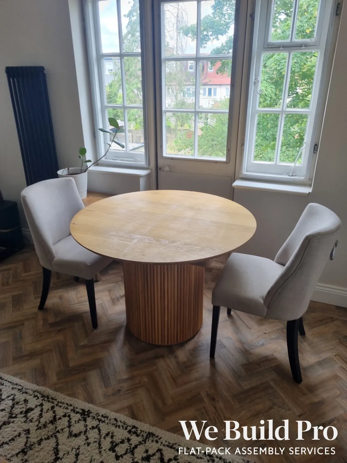 Round wooden dining table with grey upholstered chairs and large windows